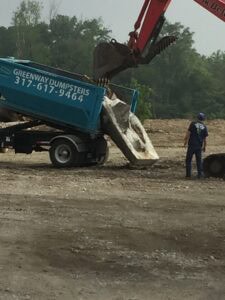 Excavator unloading concrete debris into a Greenway Dumpsters roll-off container on a construction site.