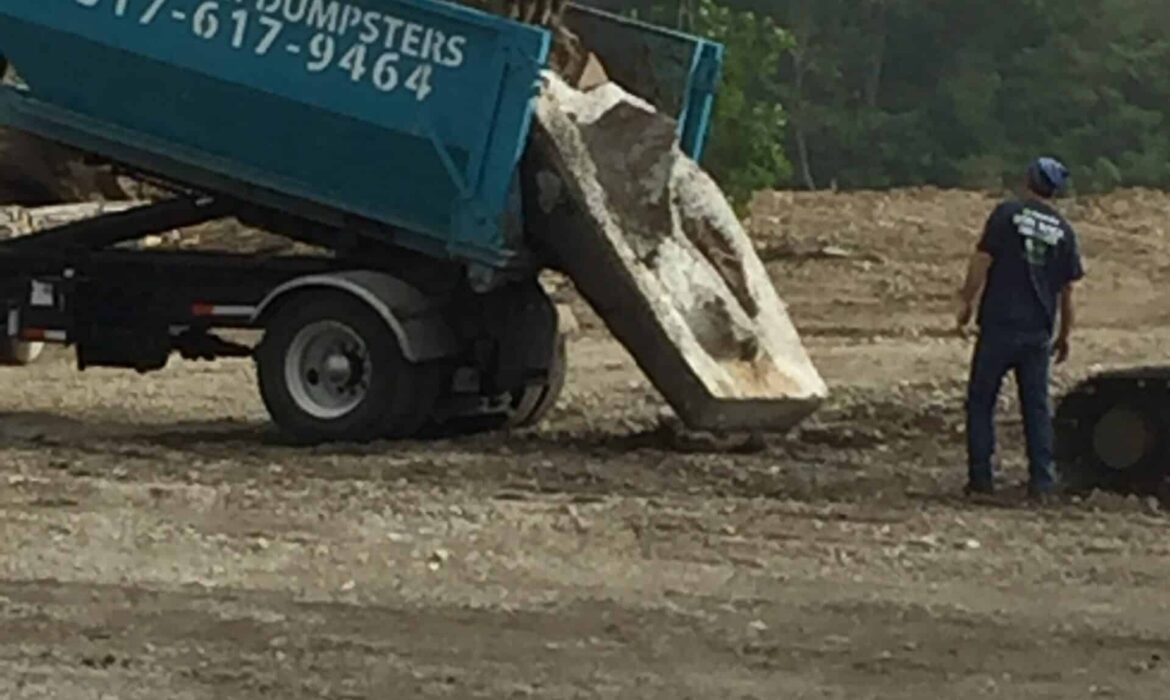 Excavator unloading concrete debris into a Greenway Dumpsters roll-off container on a construction site.