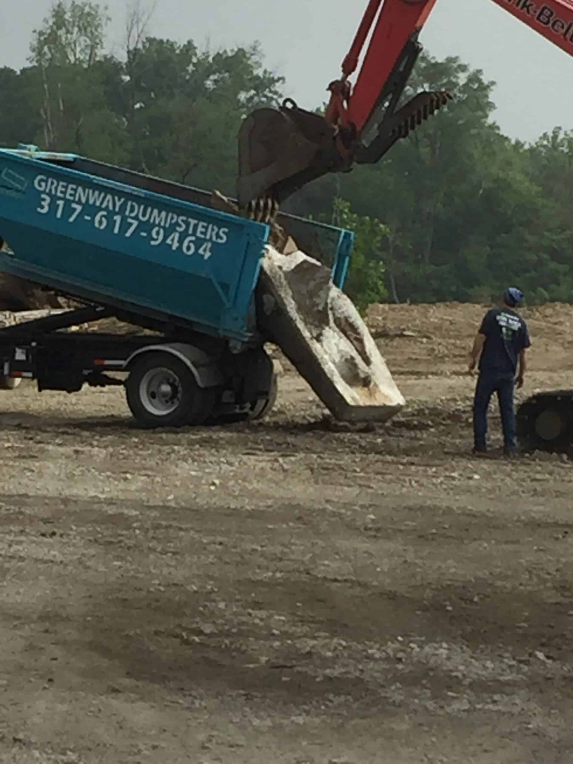 Excavator unloading concrete debris into a Greenway Dumpsters roll-off container on a construction site.